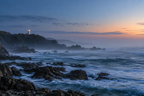 A white lighthouse overlooks dark coastal rocks, breaking surf, and blue-hour sea mist in a travel-shot concept.